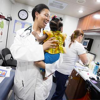 在检查室里, 一个微笑的医学院学生，脖子上挂着听诊器，抱着一个婴儿